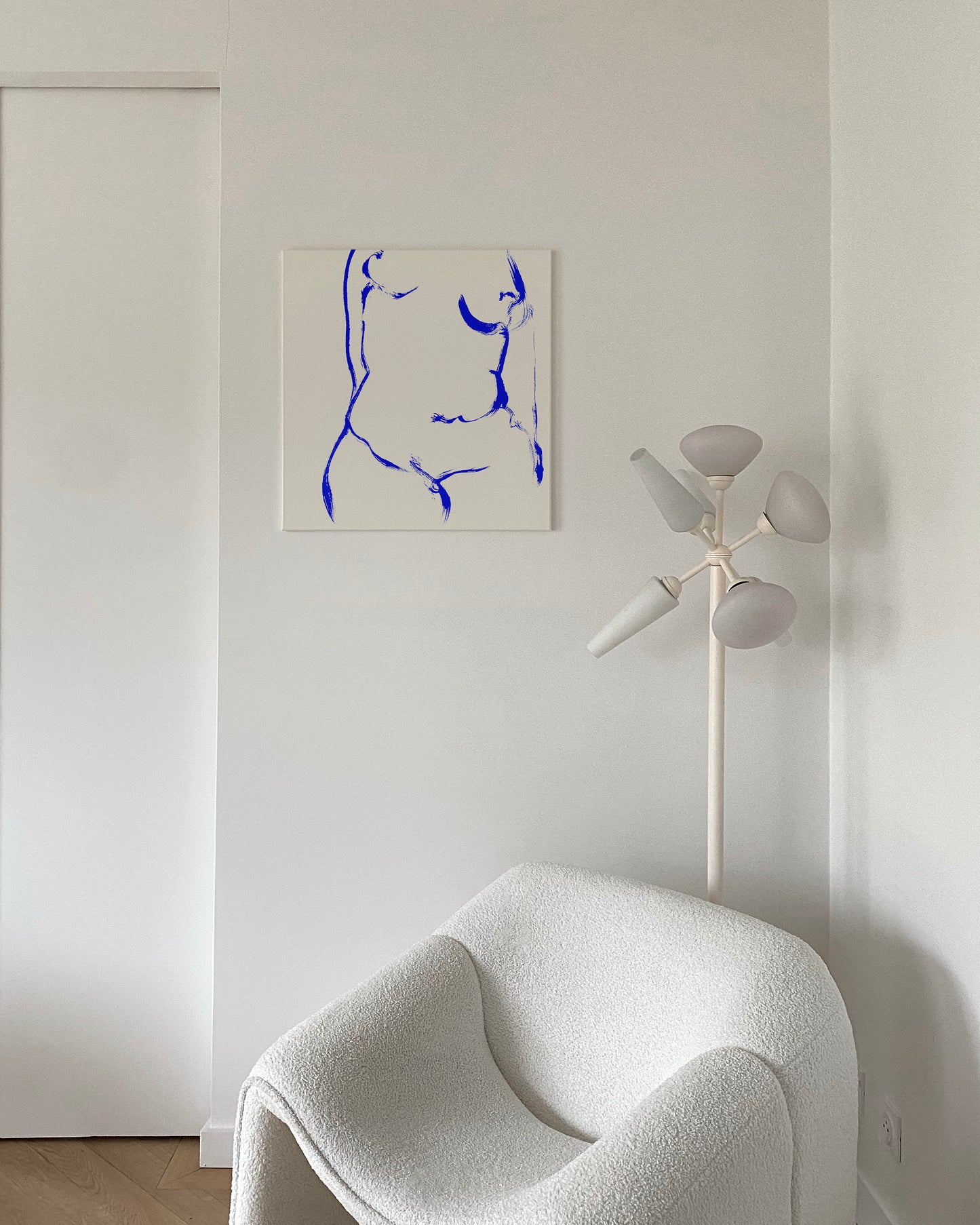 A minimalist interior scene featuring a square canvas print with a blue abstract line drawing on a white background, hung on a white wall above a white boucle armchair and next to a white multi-light floor lamp. abstract painting, acrylic on canvas, contemporary collectible wall art, anna pepe, forn studio