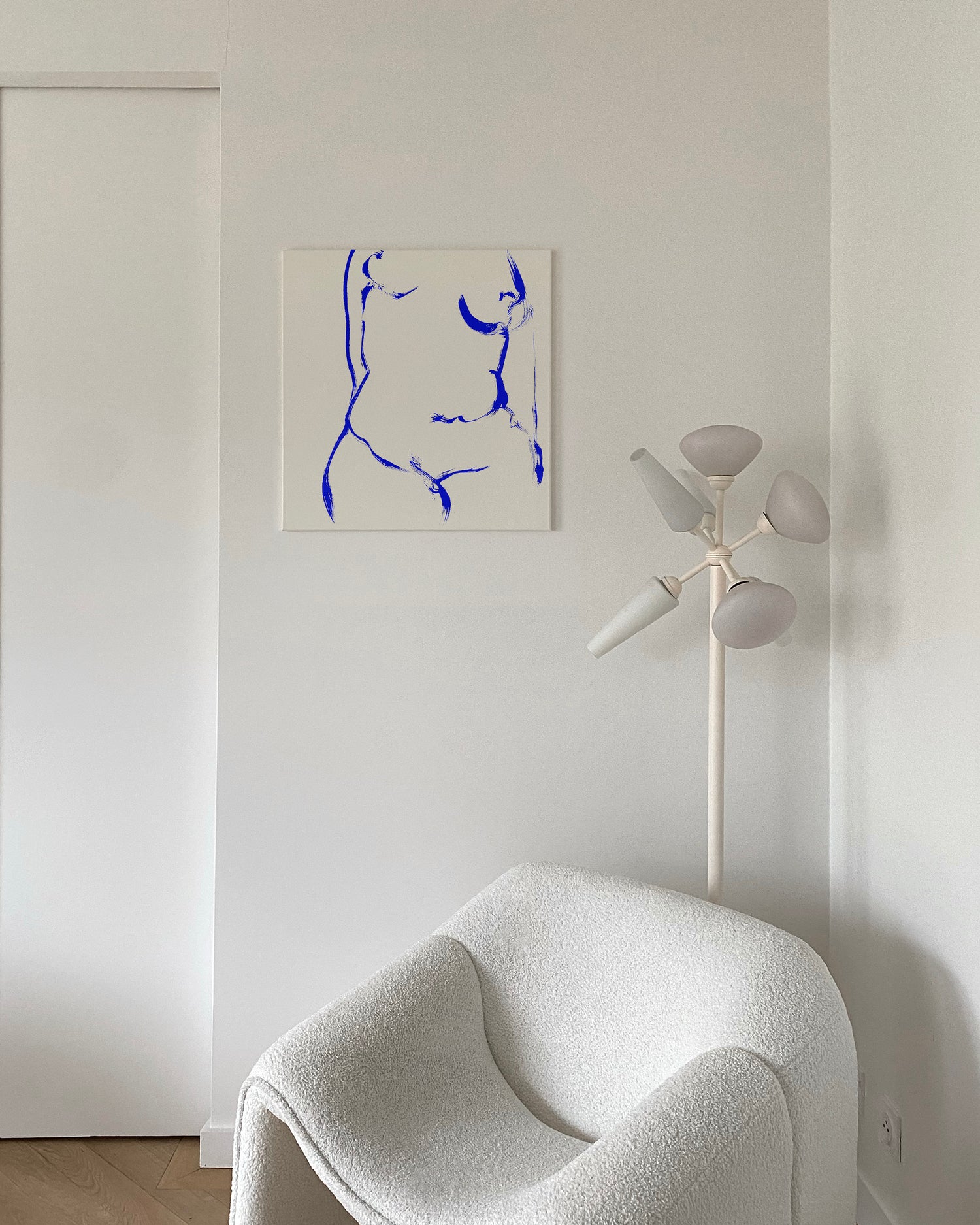 A minimalist interior scene featuring a square canvas print with a blue abstract line drawing on a white background, hung on a white wall above a white boucle armchair and next to a white multi-light floor lamp. abstract painting, acrylic on canvas, contemporary collectible wall art, anna pepe, forn studio