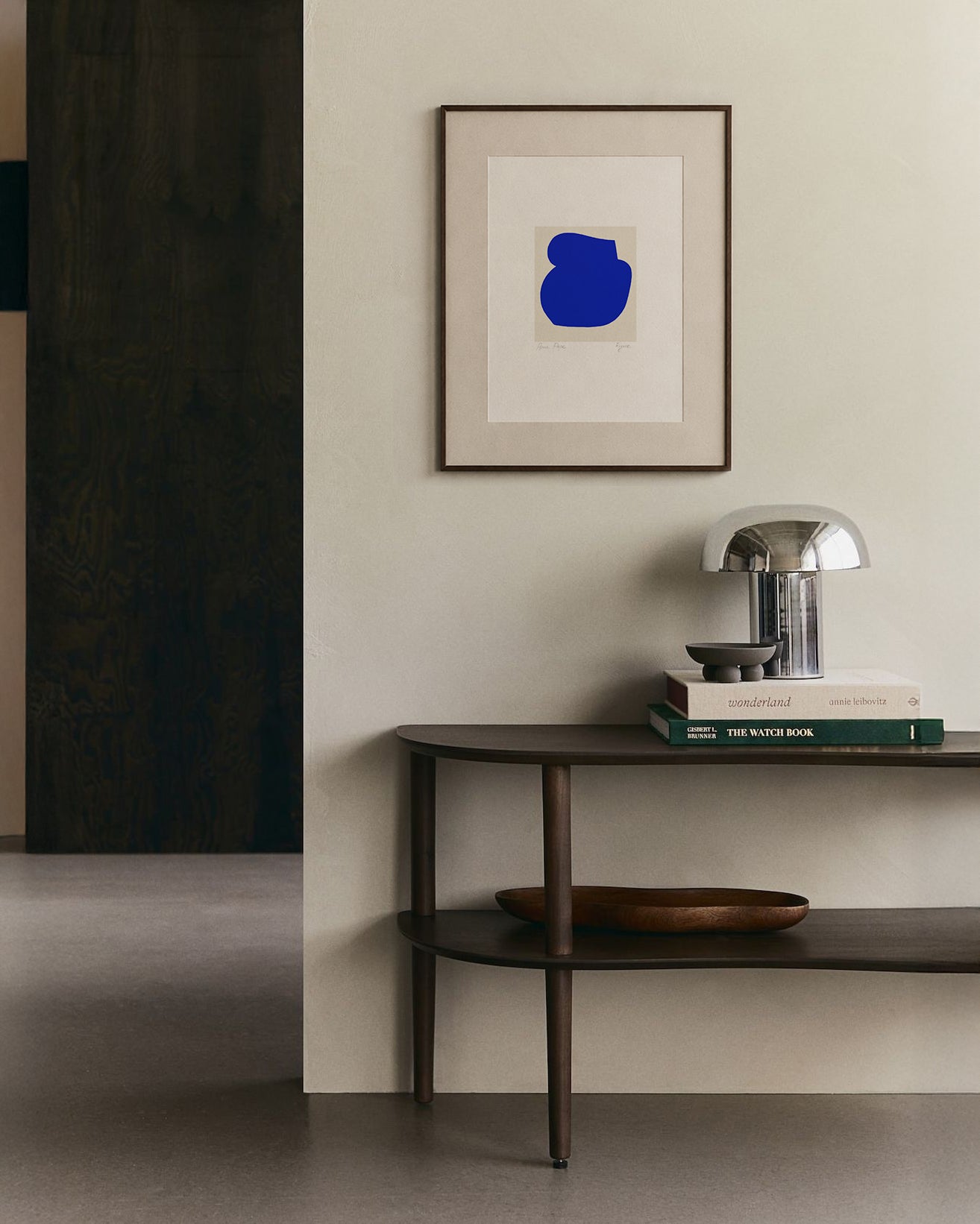 Modern interior with a dark wooden console table, books, and a lamp against a light wall with a framed abstract art piece. artist anna pepe, forn studio, abstract painting, small art work, female abstract form