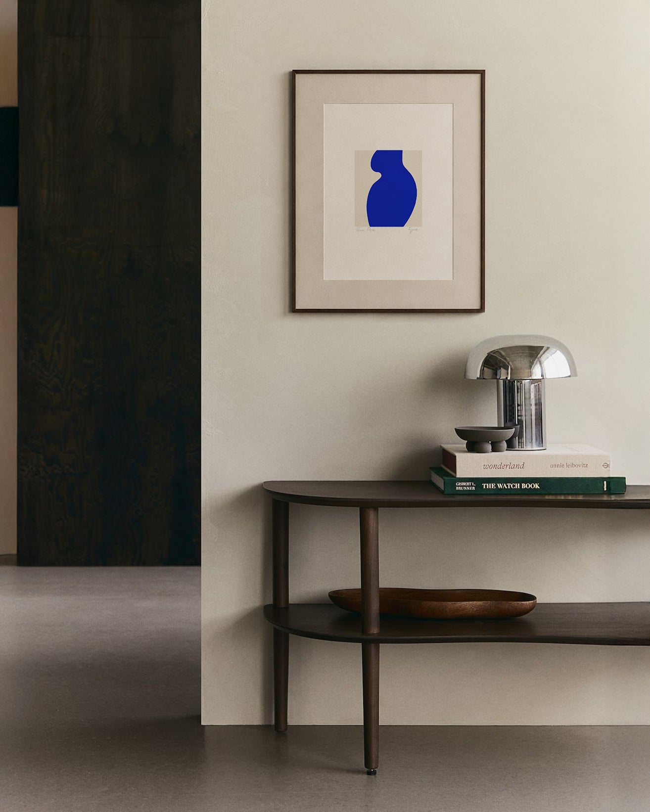 Modern interior with a dark wooden console table, books, and a lamp against a light wall with a framed abstract art piece. artist anna pepe, forn studio, abstract painting, small art work, female abstract form