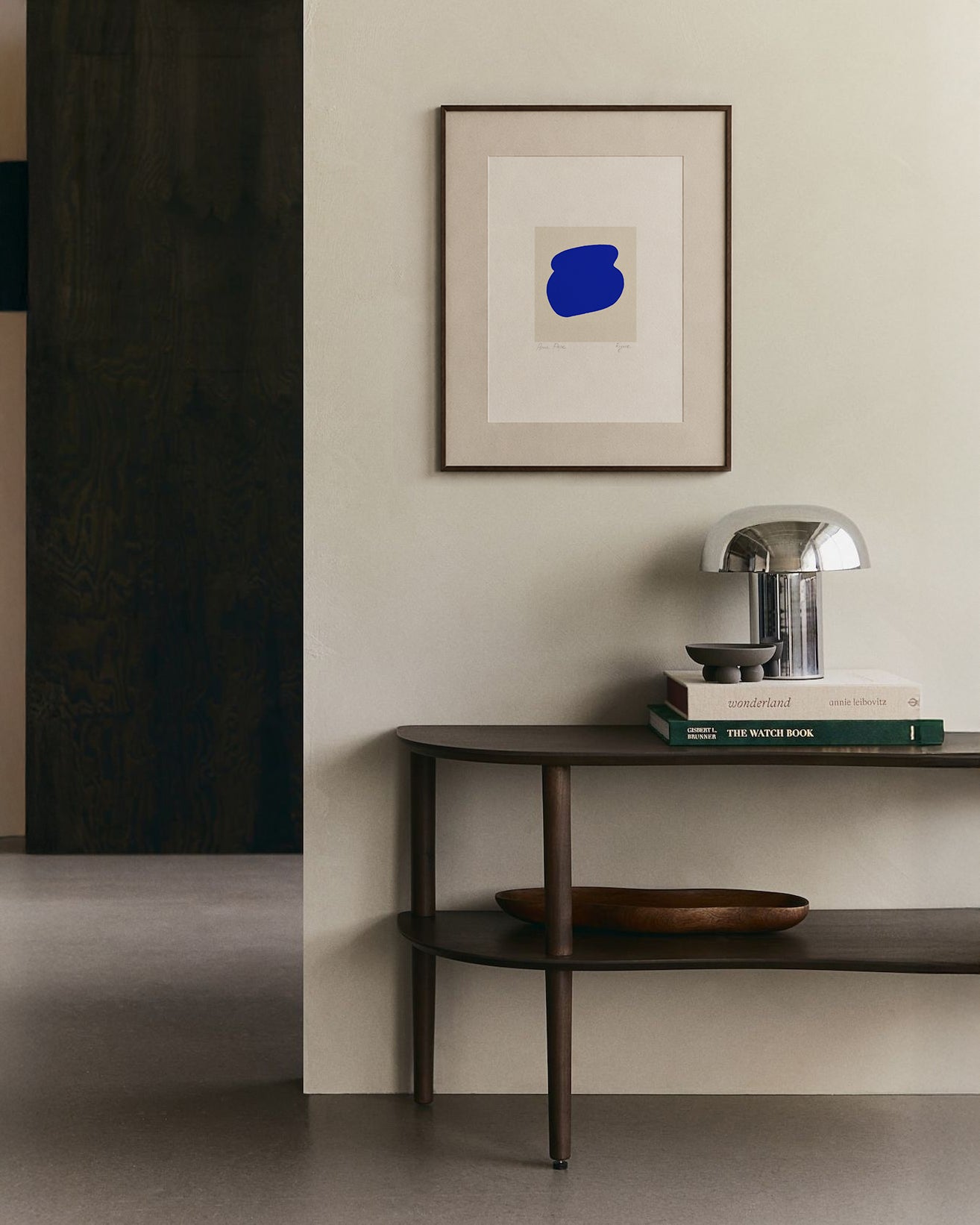 Modern interior with a dark wooden console table, books, and a lamp against a light wall with a framed abstract painting. artist anna pepe, forn studio, abstract painting, small art work, female abstract form