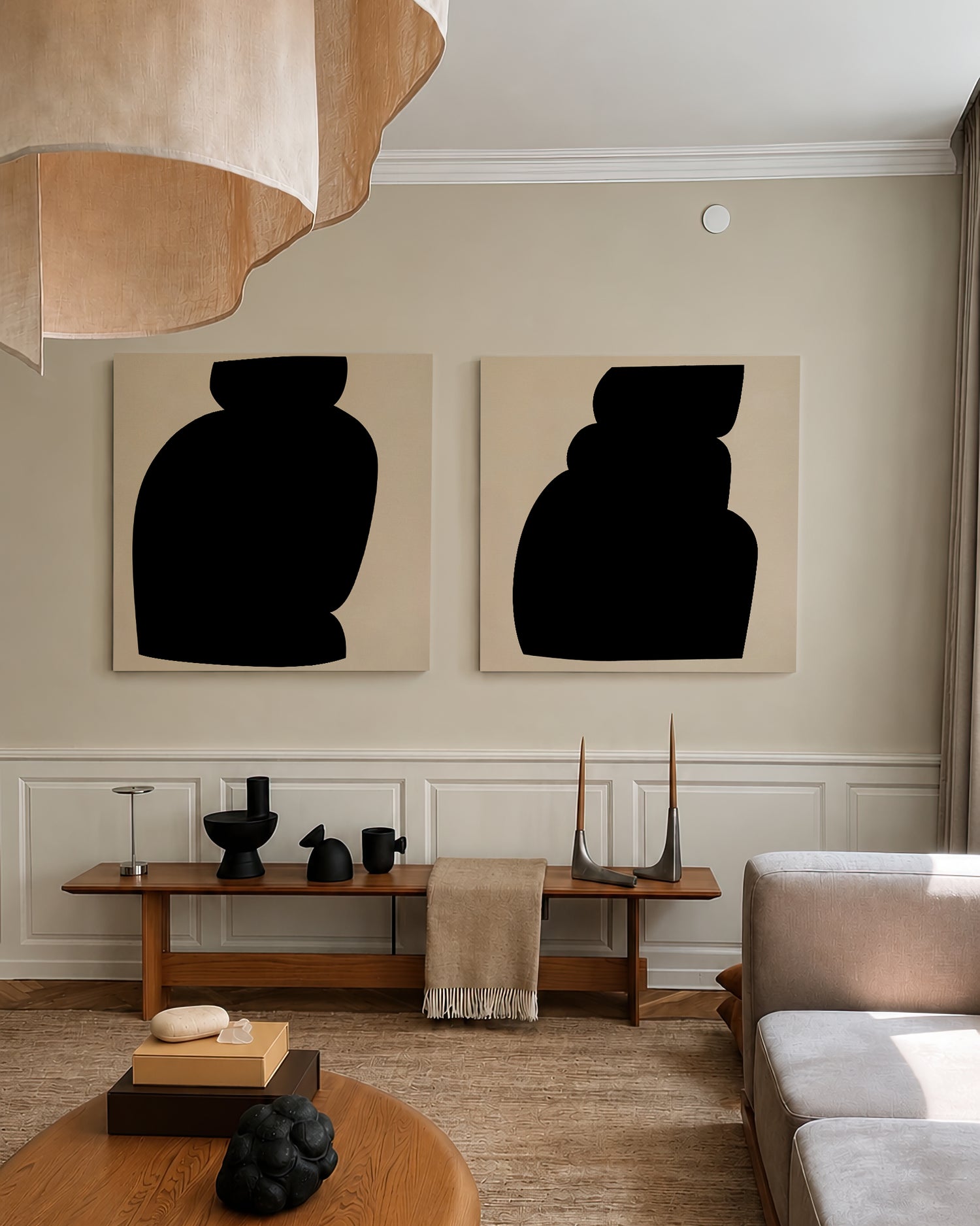 Two abstract black vase-like wall art prints with cream backgrounds hang on a beige wall above a wooden console table in a styled living room with a sofa, rug, and pendant light. large canvas, acrylic on canvas, oversized wall art, abstract art, Anna Pepe, forn Studio