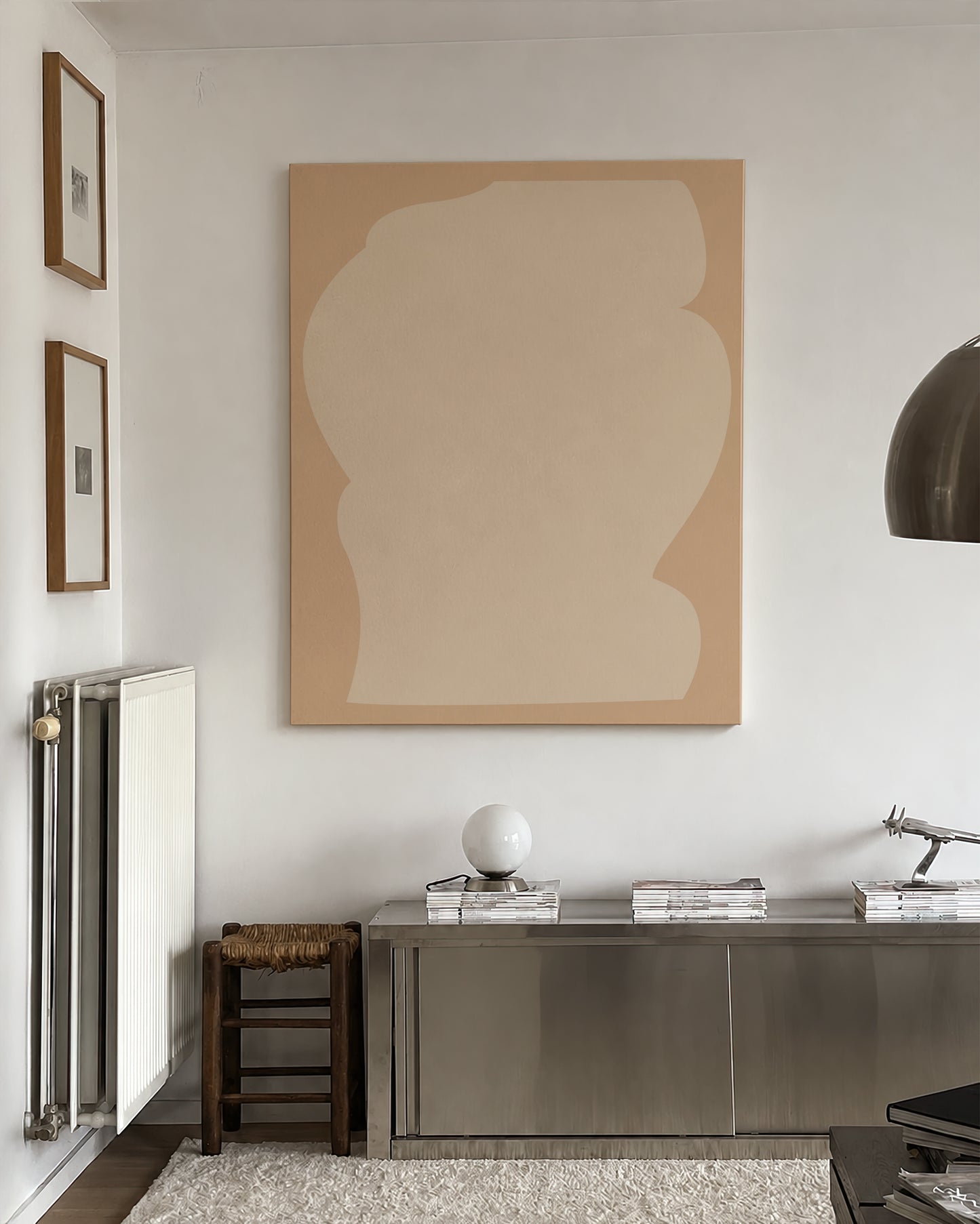 A large abstract canvas print with two beige shapes hangs on a white wall above a metallic console table in a modern, minimalist room. large canvas, acrylic on canvas, oversized wall art, abstract art, Anna Pepe, forn Studio