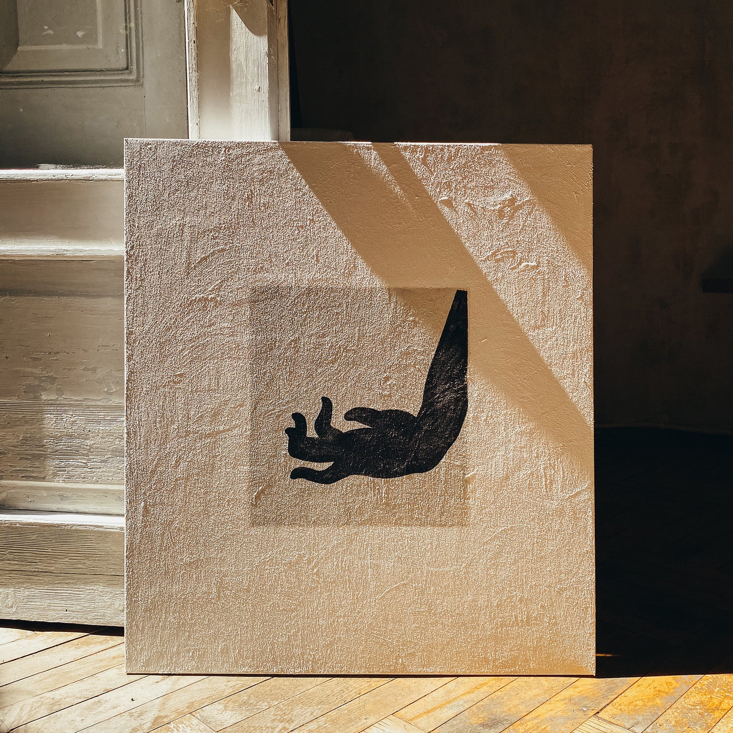A square beige canvas print with a black hand silhouette centered on a textured background, leaning against a rustic wooden wall on a wood floor. large canvas, acrylic on canvas, oversized wall art, abstract art, Anna Pepe, forn Studio